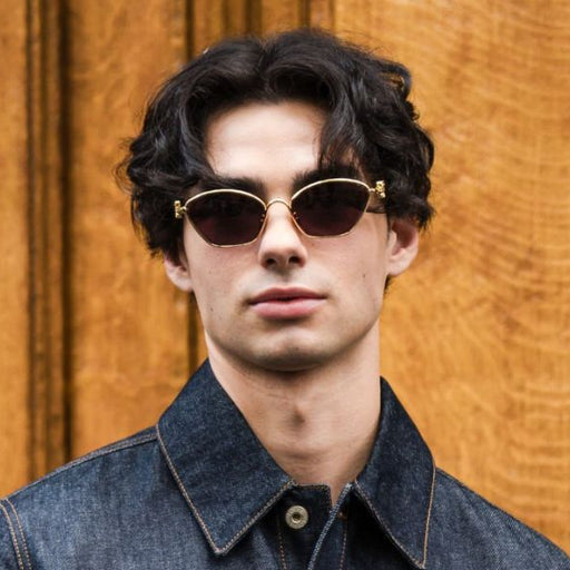 A man with dark curly hair wearing round gold-rimmed sunglasses and a denim shirt, standing in front of a warm wood-paneled background.