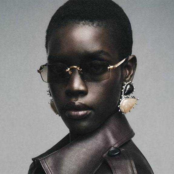A close-up portrait of a model with short dark hair wearing gold-framed rimless sunglasses, large statement earrings, and a dark brown leather jacket against a neutral gray background.