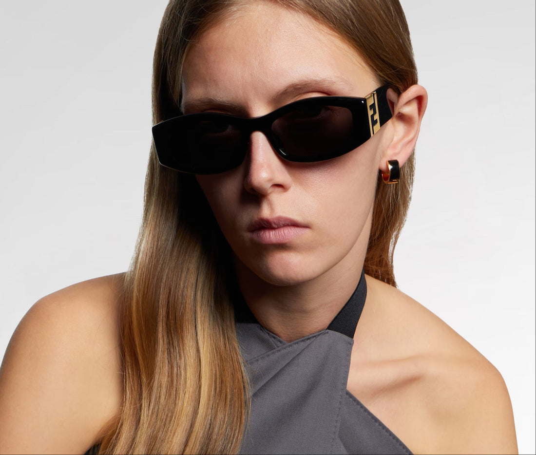 A close-up of a woman wearing black Fendi sunglasses with gold FF logos on the temples, paired with a gray halter top against a light gray background.